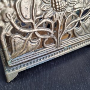 May include: A gold metal napkin holder with a sunflower design. The holder has a decorative border around the top and bottom.