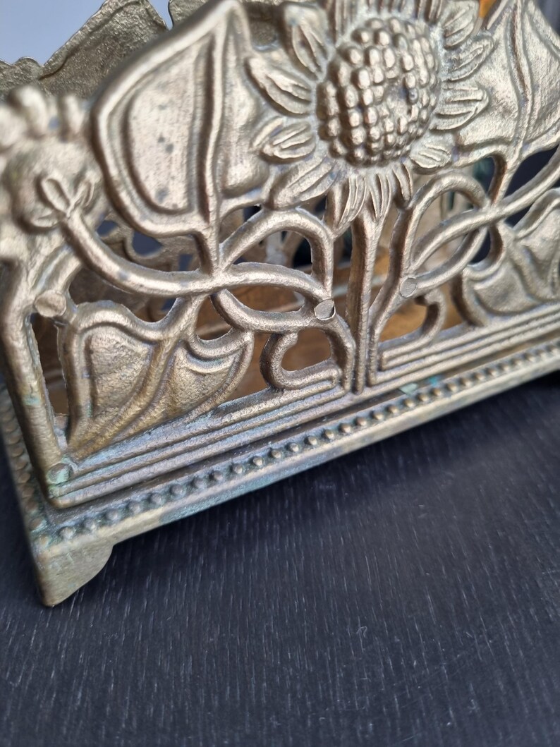 May include: A gold metal napkin holder with a sunflower design. The holder has a decorative border around the top and bottom.