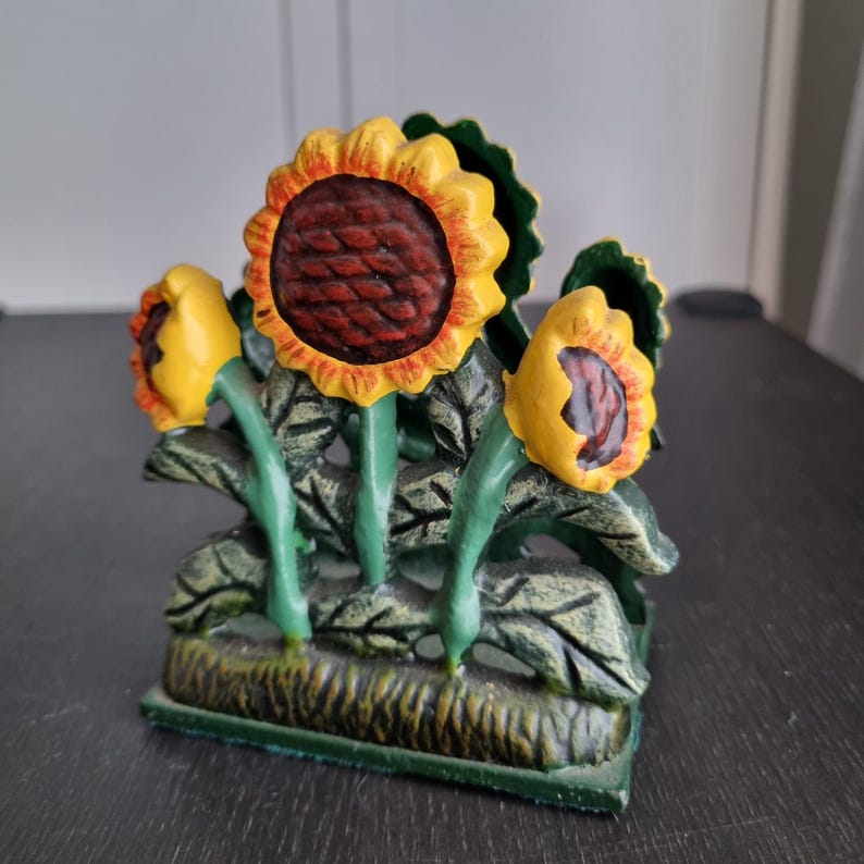 May include: A cast iron sunflower napkin holder with a green base and yellow and red sunflower accents.