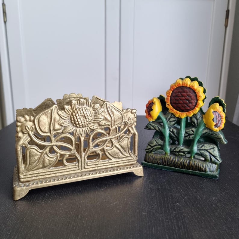 May include: Two gold-tone metal napkin holders. One is a rectangular holder with a sunflower design. The other is a smaller holder with three sunflowers and green leaves.