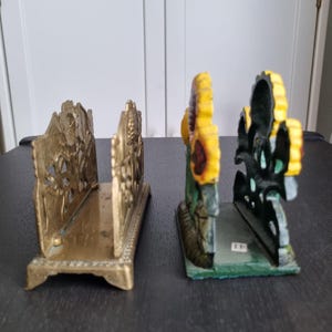May include: Two decorative napkin holders. One is made of brass with an intricate design and the other is made of cast iron with a sunflower design painted in yellow, green, and brown.