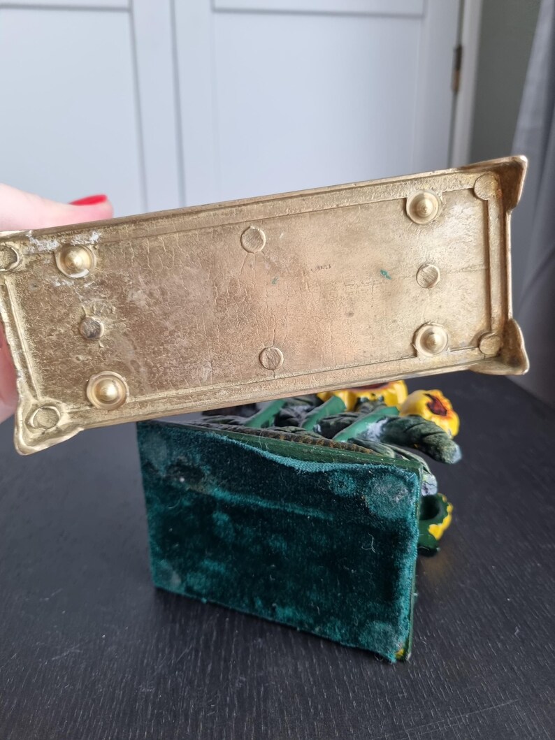 May include: A gold metal bookend with a green velvet base. The bookend features a sculpted design of sunflowers.