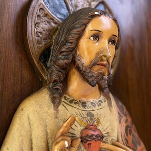 Puede incluir: Estatua detallada de una figura religiosa con cabello castaño largo y barba. La figura viste una túnica color crema y señala un corazón rojo con llamas. Un halo está detrás de la cabeza de la figura, sobre un fondo de madera.