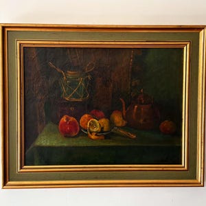 Op de afbeelding: Een olieverfschilderij in een goudkleurige en groene lijst met een stilleven. De compositie toont een koperen theepot, een glazen pot omwikkeld met touw en diverse vruchten, waaronder een geschilde sinaasappel, op een groen oppervlak.