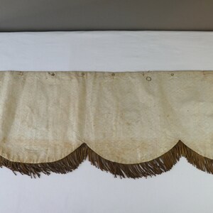 May include: A cream-colored fabric valance with a scalloped edge and brown fringe. The valance has several small metal rings along the top edge.