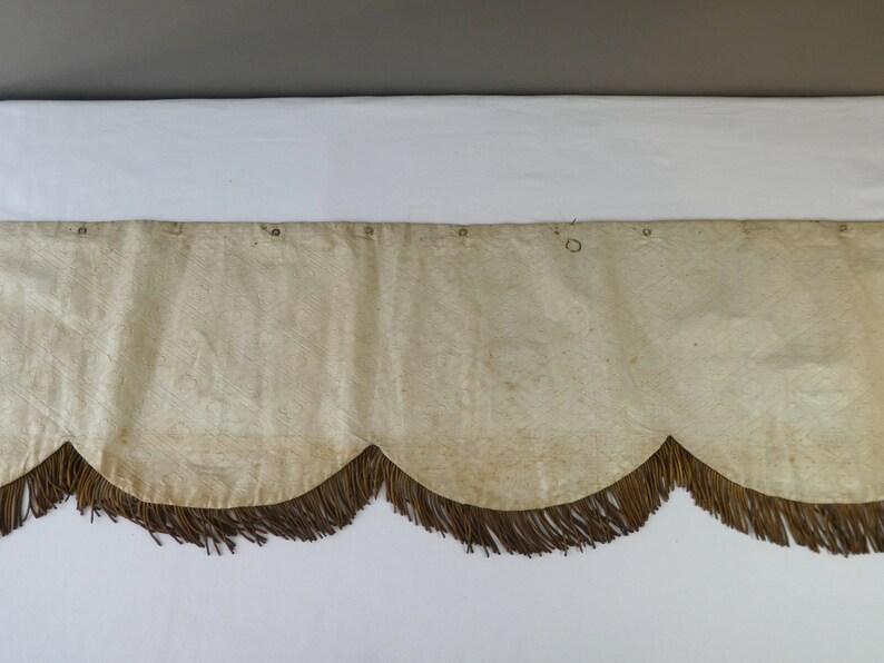 May include: A cream-colored fabric valance with a scalloped edge and brown fringe. The valance has several small metal rings along the top edge.