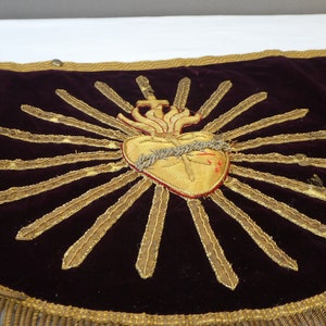 May include: A dark purple velvet cushion with a gold embroidered image of a heart with a crown of thorns and a cross. The heart is surrounded by a golden sunburst.