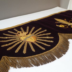 May include: A burgundy velvet valance with gold embroidered details. The center features a gold heart with flames and a crown of thorns. The right side has a gold fleur-de-lis. The valance has a gold fringe trim.