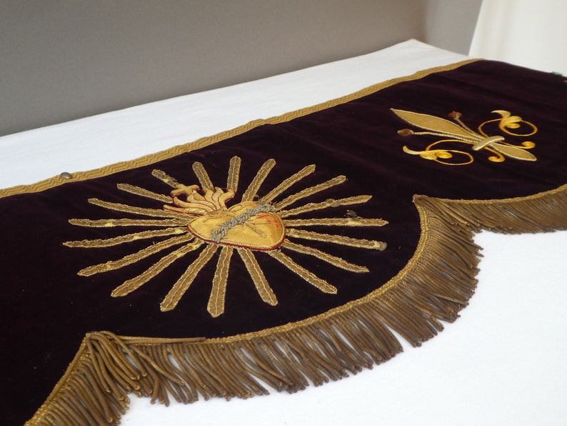 May include: A burgundy velvet valance with gold embroidered details. The center features a gold heart with flames and a crown of thorns. The right side has a gold fleur-de-lis. The valance has a gold fringe trim.