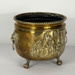 Peut inclure: Un pot en laiton vintage avec une scène en relief détaillée et des poignées en tête de lion. Le pot à trois pieds a une garniture décorative en forme de corde autour du bord. L'intérieur est noir. Une pièce décorative pour la décoration intérieure.