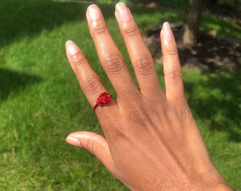 May include: A red wire rose ring. The ring is worn on the ring finger of a person's hand.
