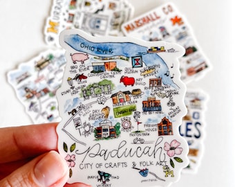 May include: A colorful illustrated map sticker of Paducah, Kentucky, with the text "City of Crafts & Folk Art". The map features landmarks like the convention center, the river, and the freight house. The sticker is white with blue, green, red, and yellow accents.