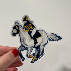 May include: A sticker featuring a watercolor illustration of a horse and jockey in a race. The horse is white with blue and yellow accents, and the jockey wears a matching uniform. The sticker is held by a hand with a red painted nail.