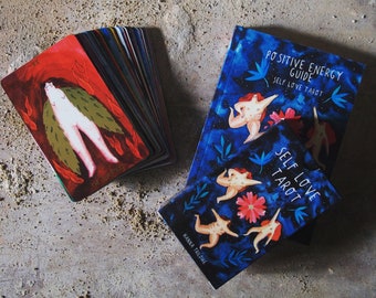 Może przedstawiać: Talia kart tarota w kolorze niebieskim i białym z tytułem "Positive Energy Guide Self Love Tarot" i podtytułem "Self Love Tarot" na okładce. Talia leży na brązowej powierzchni.