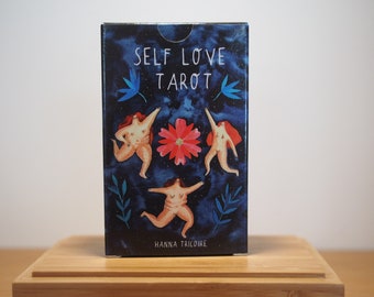May include: A box of Self Love Tarot cards with a blue background and three figures in a circle around a pink flower. The text on the box reads "Self Love Tarot" and "Hanna Tricolore".