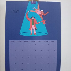 May include: A blue calendar with the word "MAY" in white text at the top. The calendar features a colorful illustration of two figures performing a magic trick under a spotlight. The calendar grid shows the days of the month, numbered 1 through 31.