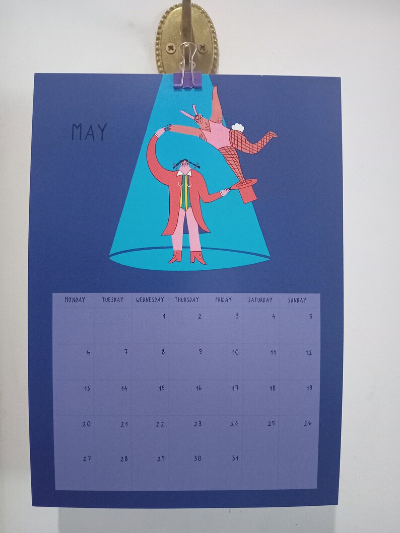 May include: A blue calendar with the word "MAY" in white text at the top. The calendar features a colorful illustration of two figures performing a magic trick under a spotlight. The calendar grid shows the days of the month, numbered 1 through 31.
