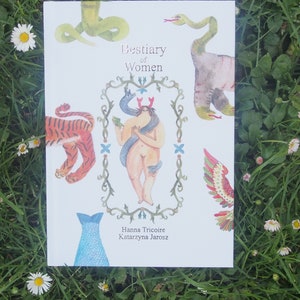 May include: A white book with the title "Bestiary of Women" and illustrations of a woman with a snake, a tiger, a fish, and a bird. The book is lying on a bed of green grass and white daisies.