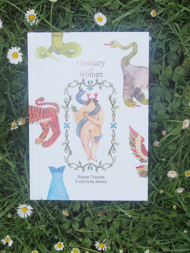 May include: A white book with the title "Bestiary of Women" and illustrations of a woman with a snake, a tiger, a fish, and a bird. The book is lying on a bed of green grass and white daisies.