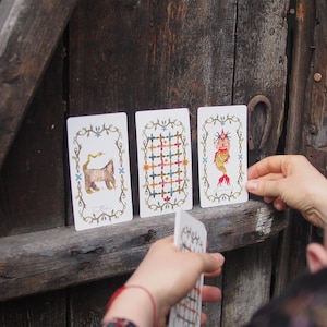 May include: Three tarot cards with illustrations on a white background. The first card shows a cat with a snake wrapped around it. The second card shows a grid pattern with different colored squares. The third card shows a mermaid with a fish tail and a crown.