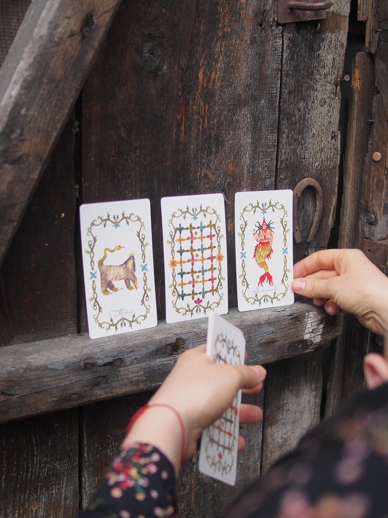 May include: Three tarot cards with illustrations on a white background. The first card shows a cat with a snake wrapped around it. The second card shows a grid pattern with different colored squares. The third card shows a mermaid with a fish tail and a crown.