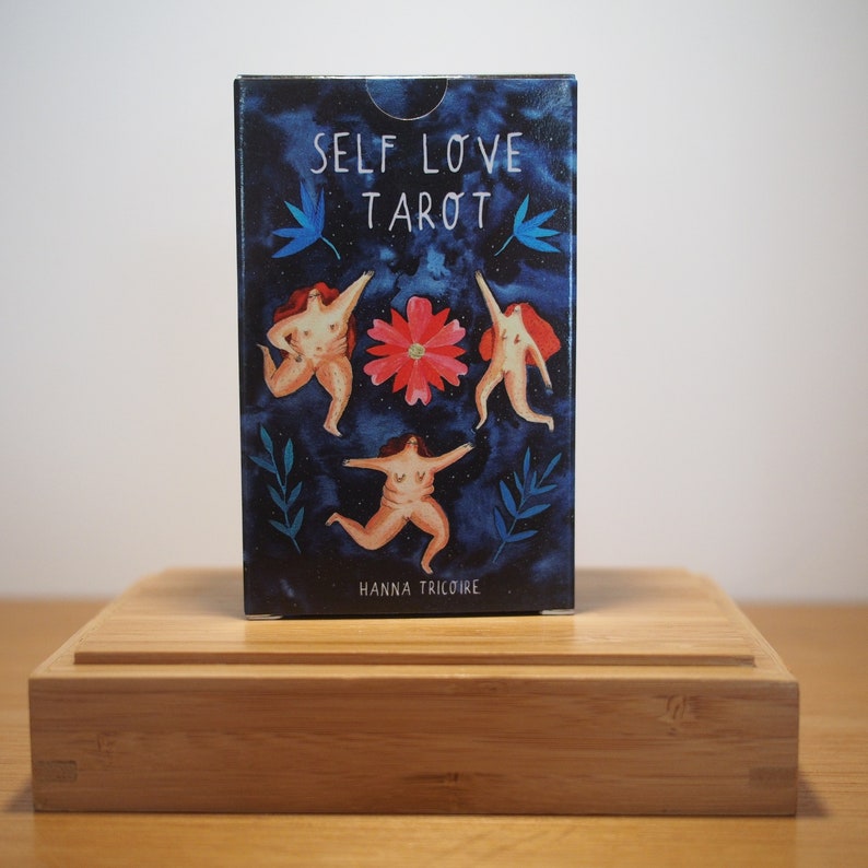 Puede incluir: Una caja de cartas de Tarot de Amor Propio con fondo azul y tres figuras en c&iacute;rculo alrededor de una flor rosa. El texto de la caja dice "Self Love Tarot" y "Hanna Tricolore".
