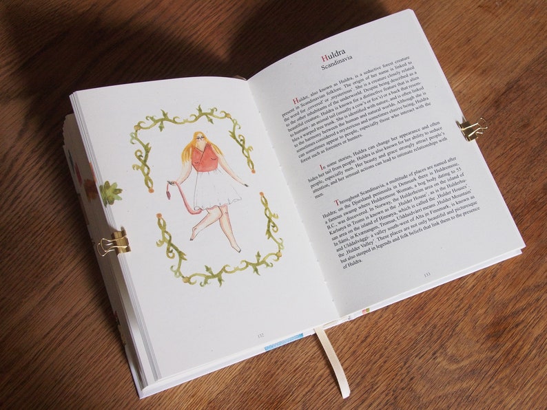 May include: An open book with a watercolor illustration of a woman with blonde hair wearing a red top and white skirt. The text on the page reads "Huldra Scandinavia".