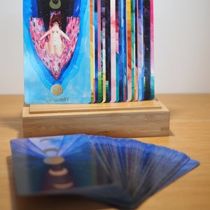 Puede incluir: Una baraja de cartas de tarot con un dise&ntilde;o abstracto y colorido. Las cartas est&aacute;n extendidas sobre una superficie de madera. La carta superior muestra a una mujer en un carro con una luna creciente sobre su cabeza. La carta est&aacute; etiquetada como "VII El Carro".