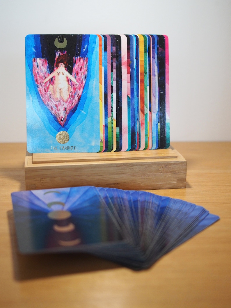 Puede incluir: Una baraja de cartas de tarot con un dise&ntilde;o abstracto y colorido. Las cartas est&aacute;n extendidas sobre una superficie de madera. La carta superior muestra a una mujer en un carro con una luna creciente sobre su cabeza. La carta est&aacute; etiquetada como "VII El Carro".