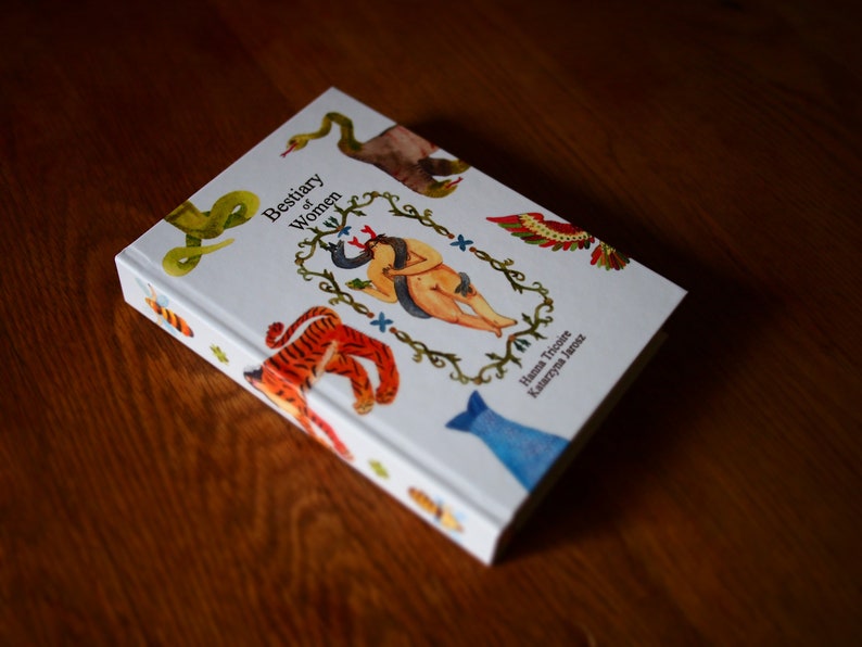 May include: A white book with the title "Bestiary of Women" and illustrations of a snake, tiger, fish, and a woman with a snake wrapped around her. The book is by Hanna Tricoche and Kateryna Yarmosh.