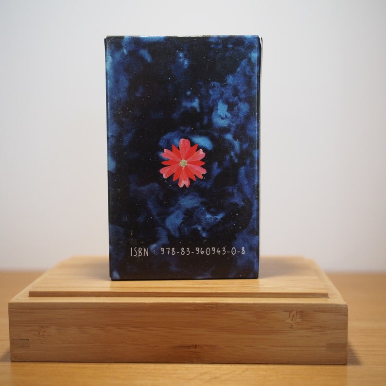 Puede incluir: Un libro azul con una flor roja en la portada. El libro tiene el texto "ISBN 978-83-960943-0-8" en la parte inferior.