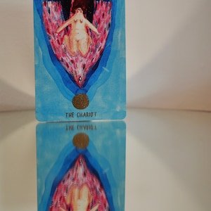 Puede incluir: La carta del tarot El Carro presenta a una mujer con una corona de flores, sentada en un carro tirado por dos criaturas aladas. El fondo es de un azul y un rosa vibrantes con una luna creciente en la esquina superior izquierda.