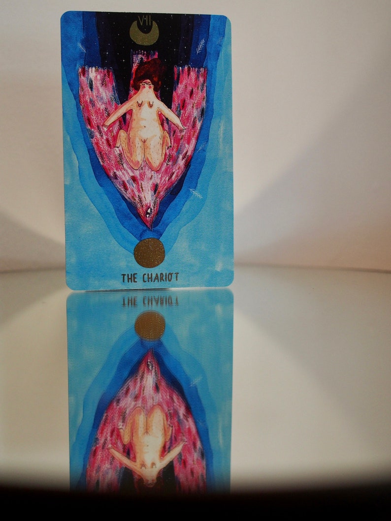 Puede incluir: La carta del tarot El Carro presenta a una mujer con una corona de flores, sentada en un carro tirado por dos criaturas aladas. El fondo es de un azul y un rosa vibrantes con una luna creciente en la esquina superior izquierda.
