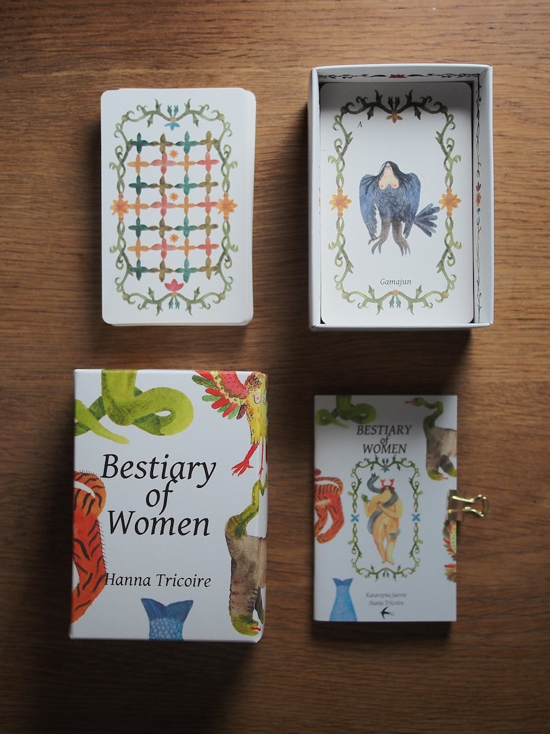 May include: A set of cards with illustrations of mythical creatures, titled "Bestiary of Women" by Hanna Tricoire. The cards are in a white box with a green, orange, and white patterned card on top. The box is on a wooden surface.