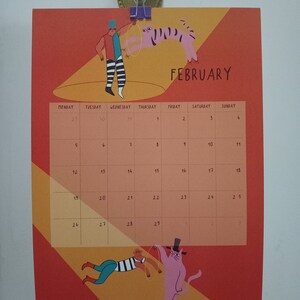 May include: A February calendar with a red and orange background. The calendar features a circus theme with illustrations of a person juggling a tiger and a person holding a cat on a string. The calendar is titled "FEBRUARY" in black text.
