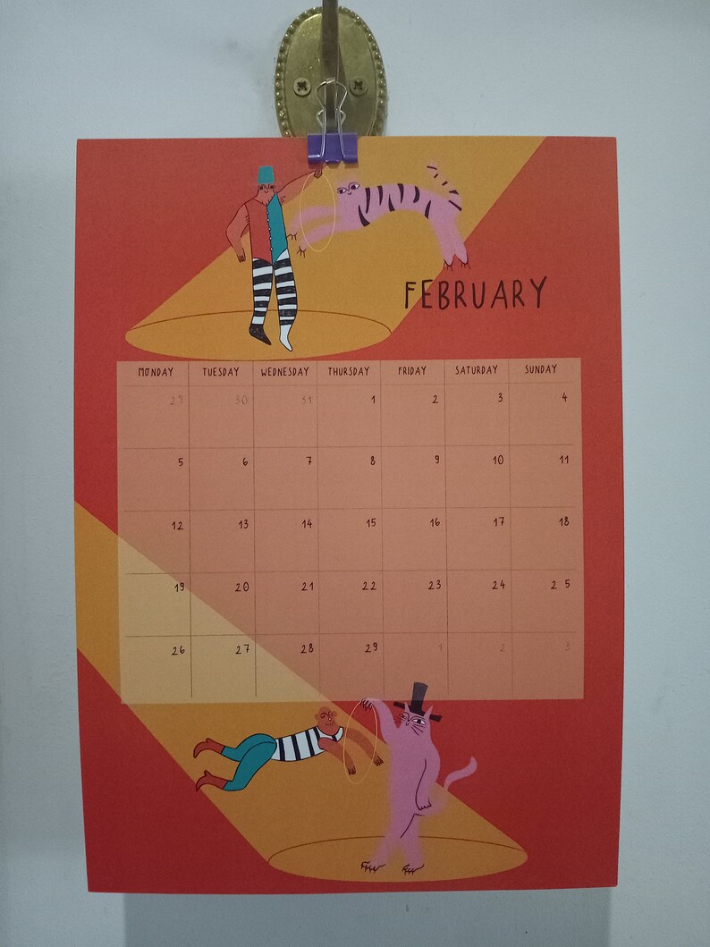 May include: A February calendar with a red and orange background. The calendar features a circus theme with illustrations of a person juggling a tiger and a person holding a cat on a string. The calendar is titled "FEBRUARY" in black text.