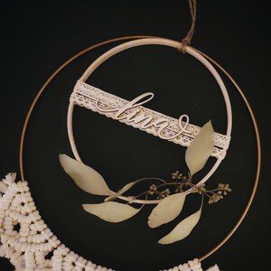 May include: A white macrame wall hanging with a wooden sign that says "love" and dried eucalyptus leaves on a gold hoop.