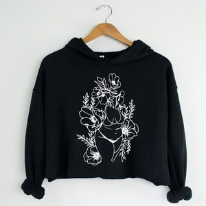 May include: Black cropped hoodie with a white outline of a woman's body surrounded by flowers.