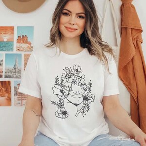 May include: White t-shirt with a black line drawing of a woman's body with flowers around her. The woman is wearing a bikini.