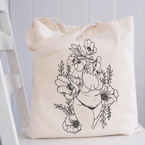 May include: A white tote bag with a black line drawing of a person with flowers growing from their head. The person is wearing a bikini and the flowers are stylized.