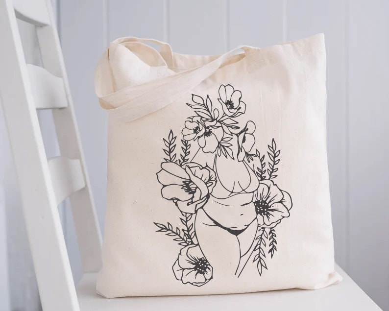 May include: A white tote bag with a black line drawing of a person with flowers growing from their head. The person is wearing a bikini and the flowers are stylized.