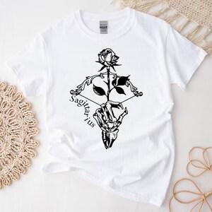 May include: White t-shirt with a black graphic of a skeletal hand holding a rose and an arrow with the word "Sagittarius" written below.