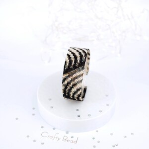 K&ouml;nnte beinhalten: Ein schwarz-wei&szlig;es Perlenarmband mit Zebra-Muster. Das Armband befindet sich auf einem wei&szlig;en Pr&auml;sentationsst&auml;nder mit dem Text "Crafty Bead" darauf.