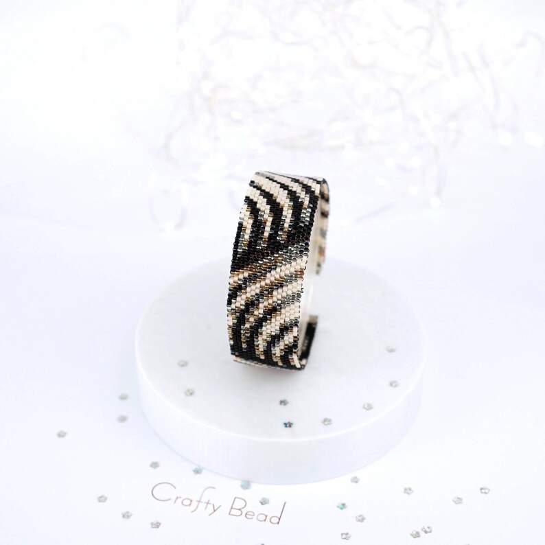 K&ouml;nnte beinhalten: Ein schwarz-wei&szlig;es Perlenarmband mit Zebra-Muster. Das Armband befindet sich auf einem wei&szlig;en Pr&auml;sentationsst&auml;nder mit dem Text "Crafty Bead" darauf.