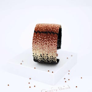 Op de afbeelding: Een kralenarmband met een gradi&euml;nt van koperen, ros&eacute;gouden en zwarte kralen. De armband is gemaakt met kleine, vierkante kralen en heeft een geometrisch patroon.