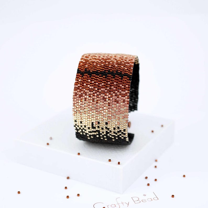 Op de afbeelding: Een kralenarmband met een gradi&euml;nt van koperen, ros&eacute;gouden en zwarte kralen. De armband is gemaakt met kleine, vierkante kralen en heeft een geometrisch patroon.