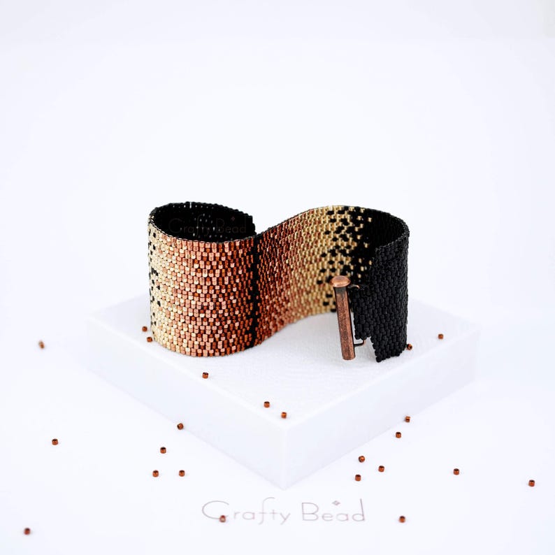 Op de afbeelding: Een armband van zwarte, koperen en gouden kralen met een koperen sluiting. De armband ligt op een wit oppervlak met kleine, verspreide koperen kralen.