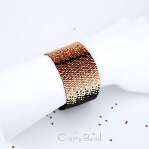 Op de afbeelding: Een kralenarmband met een verloop van koperen, ros&eacute;gouden en zwarte kralen. De armband ligt op een wit oppervlak met de tekst "Crafty Bead" in zwart.