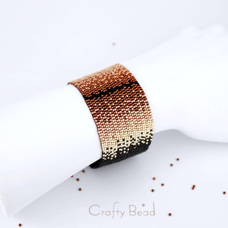 Op de afbeelding: Een kralenarmband met een verloop van koperen, ros&eacute;gouden en zwarte kralen. De armband ligt op een wit oppervlak met de tekst "Crafty Bead" in zwart.