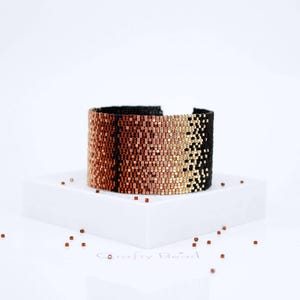 Op de afbeelding: Een zwarte, koperen en gouden kralen armband met een geometrisch patroon. De armband ligt op een wit oppervlak met kleine koperen kralen eromheen.